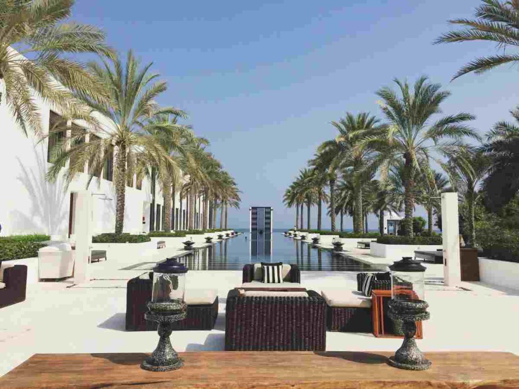 Palm-lined hotel swimming pool with lounge seating and lanterns under a clear blue sky in Oman